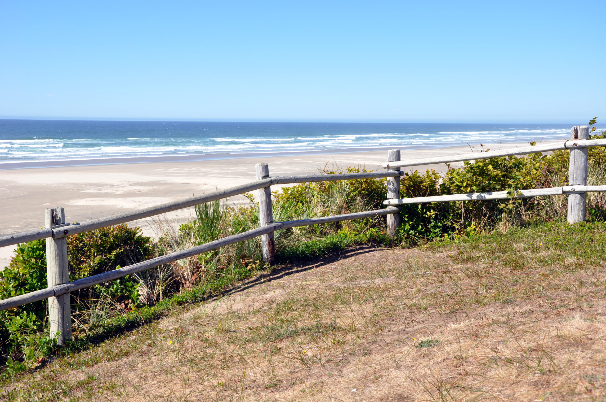 A broad Oregon beach with surf beyond a fence and driftwood near the shoreline