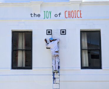 A person on a ladder paints a large QR code onto a white wall.