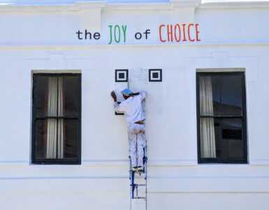 A person on a ladder paints a large QR code onto a white wall.