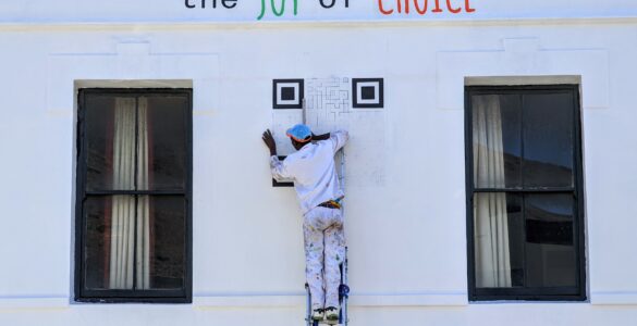 A person on a ladder paints a large QR code onto a white wall.
