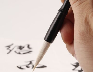 A hand uses a calligraphy brush to write Japanese characters on paper.