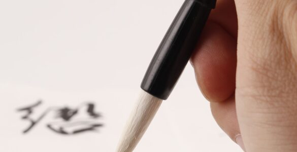 A hand uses a calligraphy brush to write Japanese characters on paper.