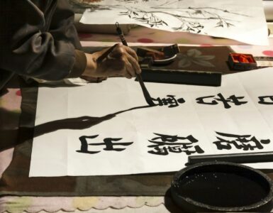 A person practices Japanese calligraphy with brush and ink on white paper.