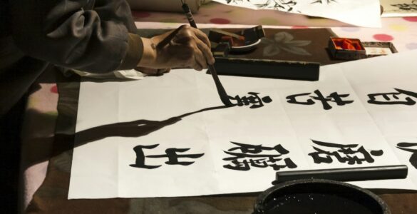 A person practices Japanese calligraphy with brush and ink on white paper.