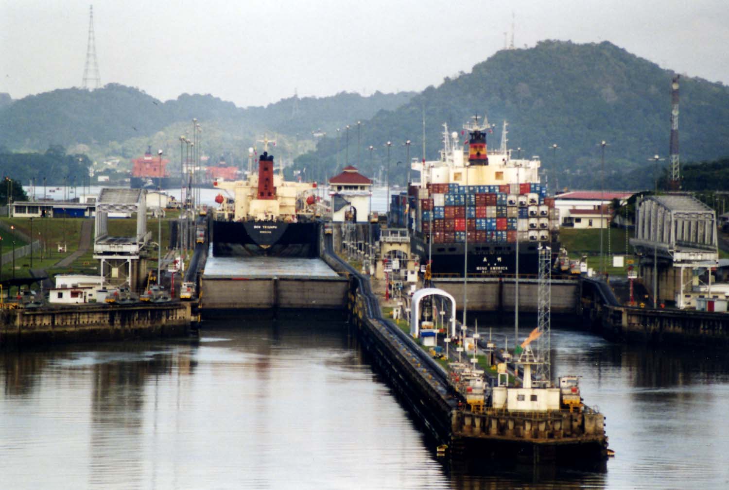 Ships move through the Miraflores Locks of the Panama Canal with forested hills behind them.
