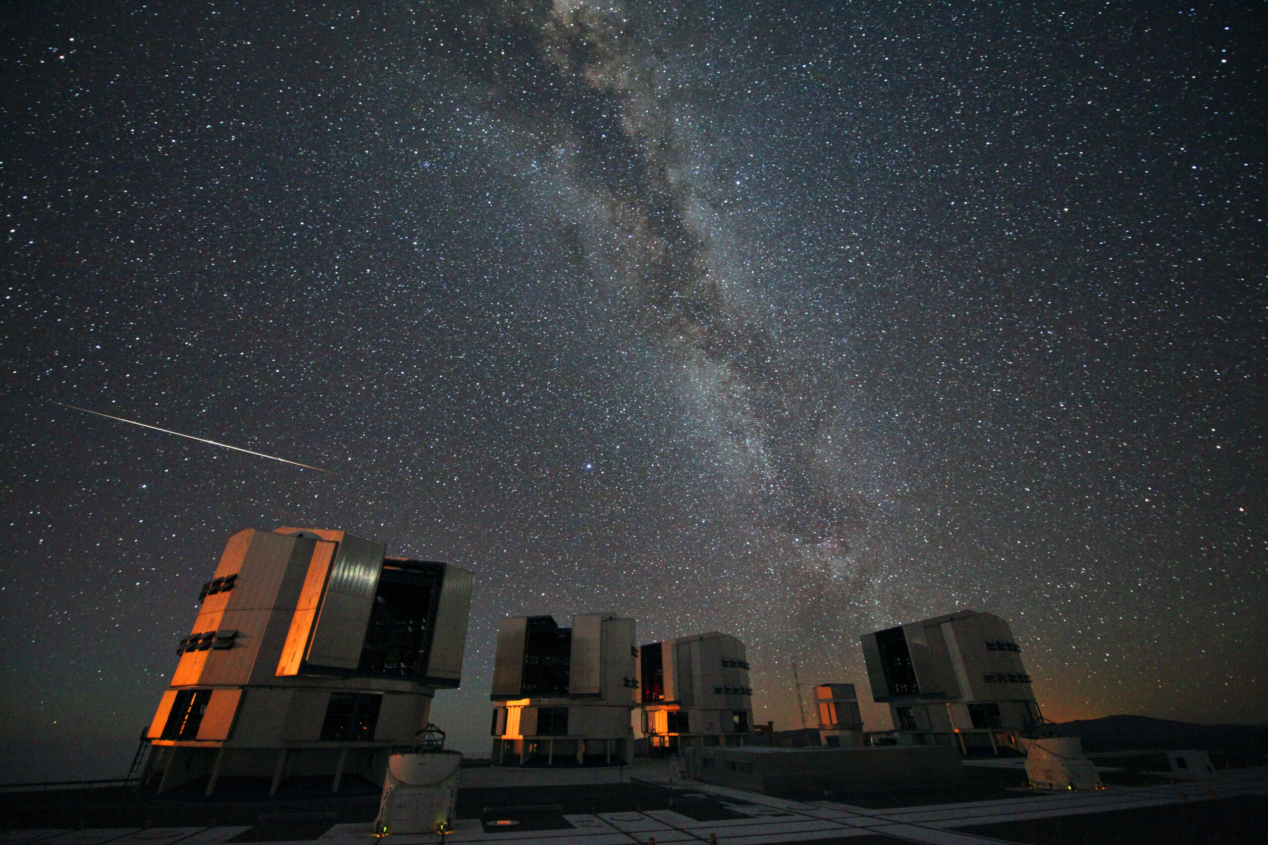 A bright Perseid meteor streaks across the Milky Way above observatory telescopes at night.