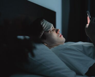 A person wearing a sleep mask checks a phone while lying in bed at night, showing how late screen use can affect sleep debt.