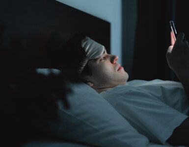 A person wearing a sleep mask checks a phone while lying in bed at night, showing how late screen use can affect sleep debt.