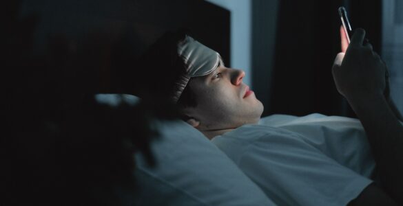 A person wearing a sleep mask checks a phone while lying in bed at night, showing how late screen use can affect sleep debt.