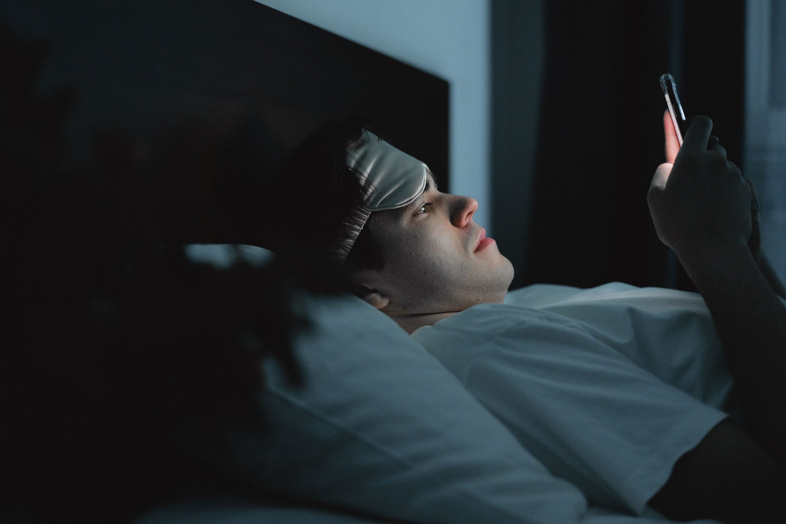 A person wearing a sleep mask checks a phone while lying in bed at night.