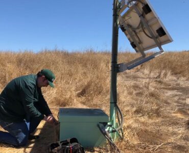 A technician services a ShakeAlert earthquake early warning sensor station in dry grassland.