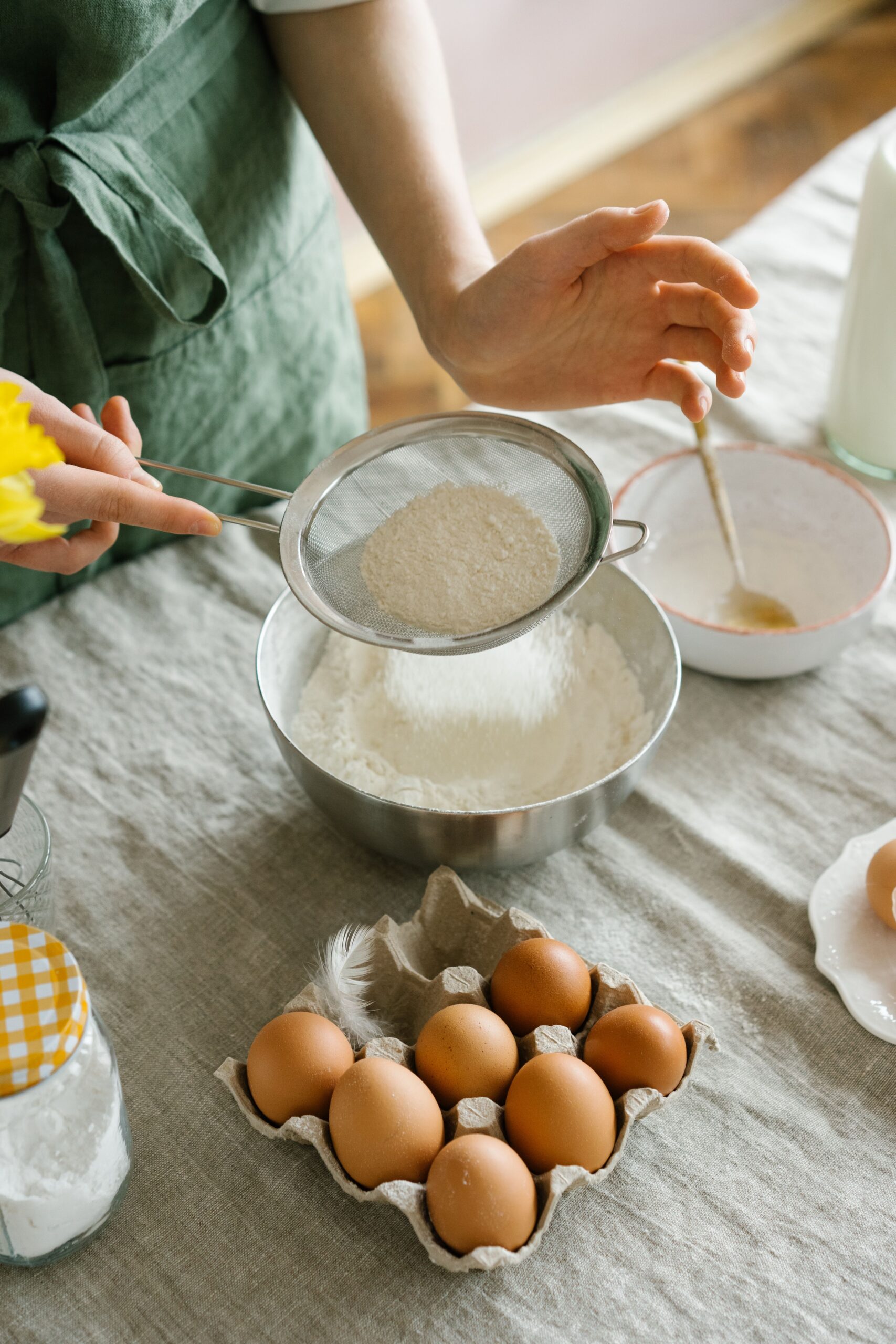 A person sifts flour into a bowl while preparing baking ingredients on a table