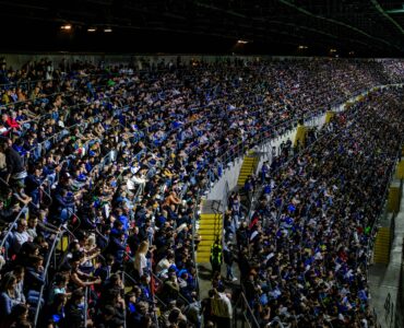 A large crowd watches a soccer match in a brightly lit stadium.