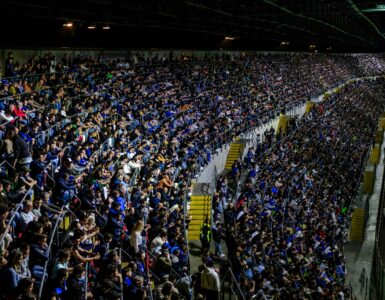 A large crowd watches a soccer match in a brightly lit stadium.