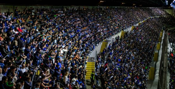 A large crowd watches a soccer match in a brightly lit stadium.
