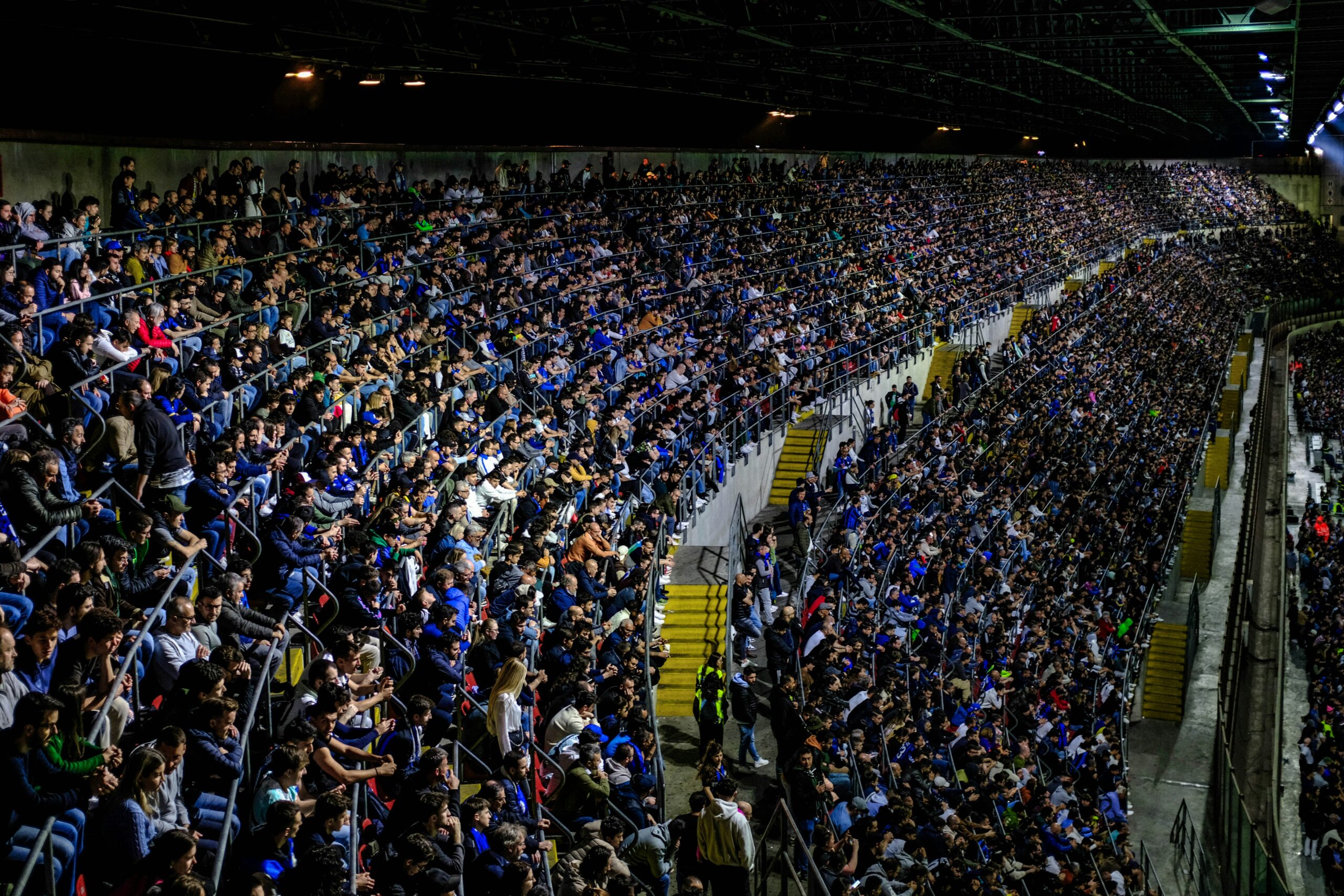 A large crowd watches a soccer match in a brightly lit stadium.