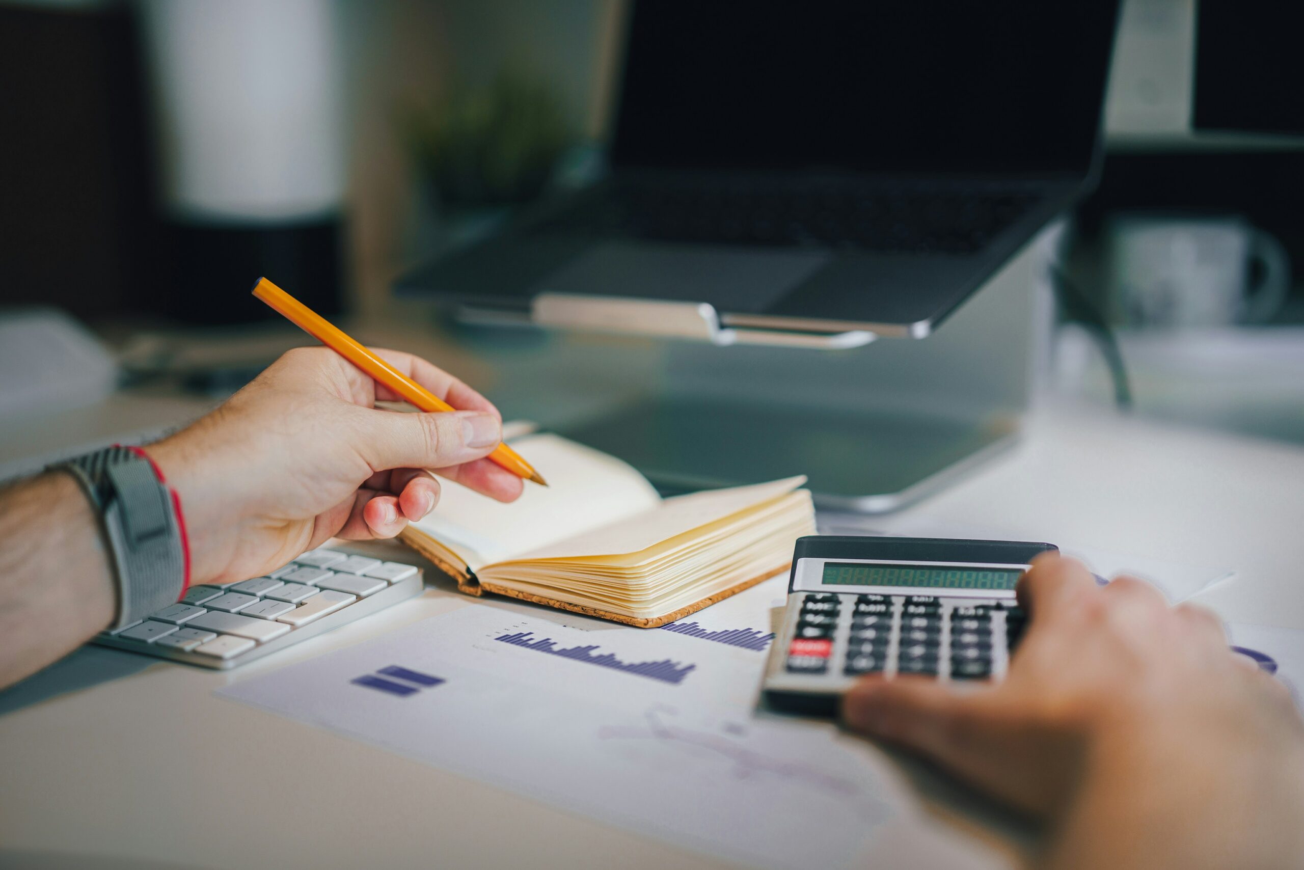 A person writes in a notebook while using a calculator for cost planning.