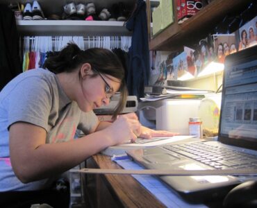 A student writing at a dorm room desk with everyday study materials nearby