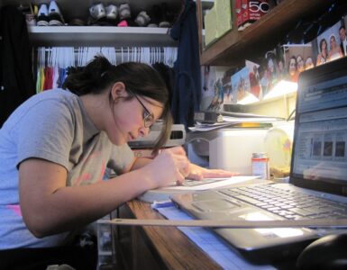 A student writing at a dorm room desk with everyday study materials nearby