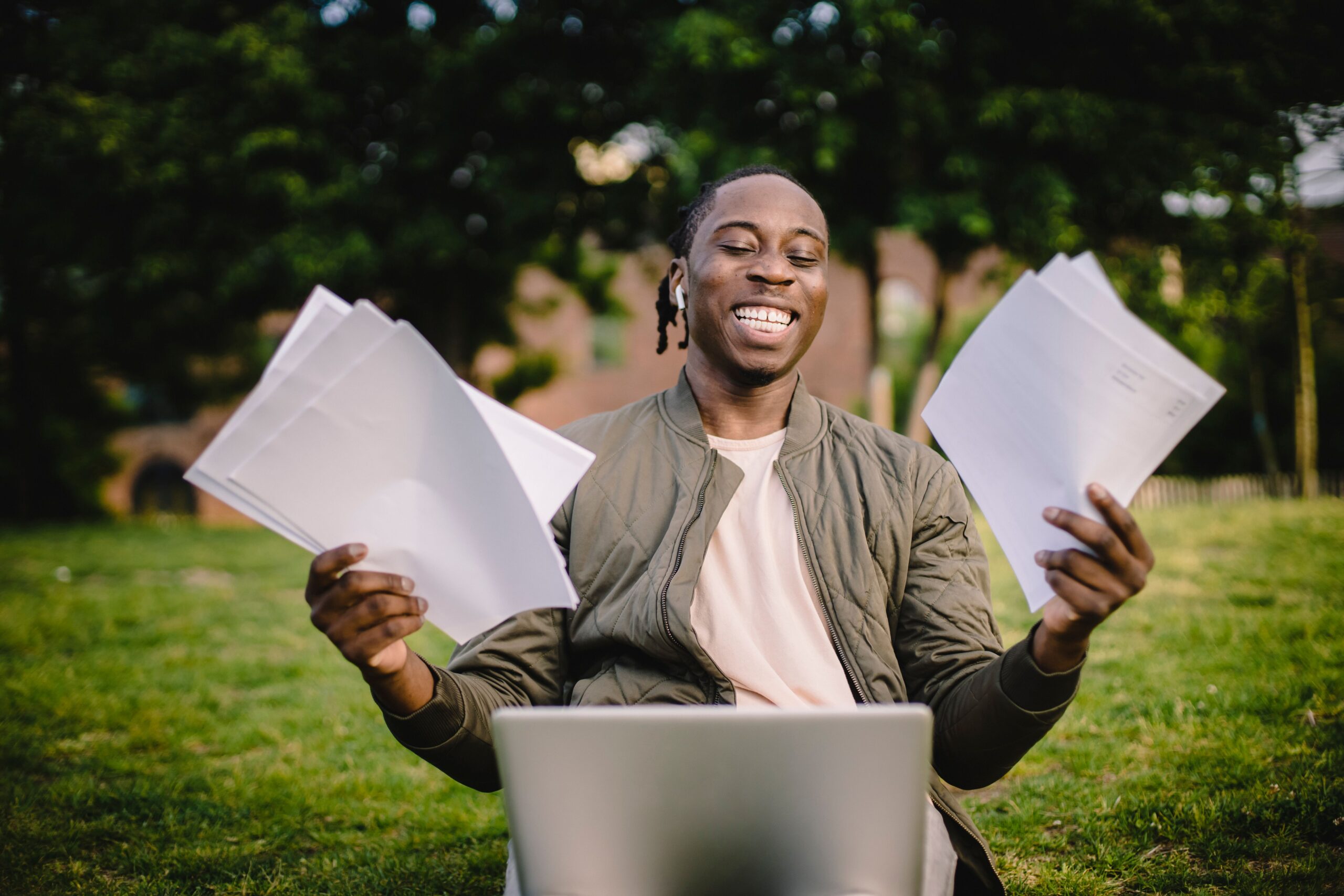 A student smiles while holding papers beside a laptop outdoors.