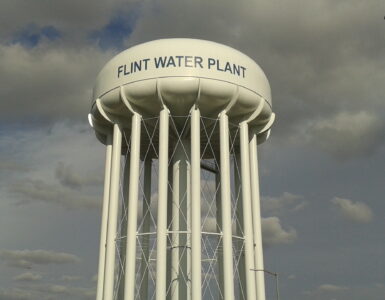 A water tower at a treatment plant rises above utility buildings and a tree-lined street.