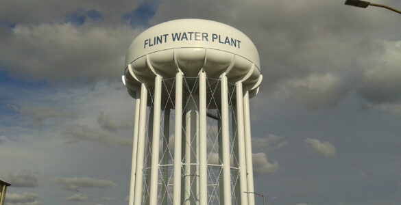 A water tower at a treatment plant rises above utility buildings and a tree-lined street.
