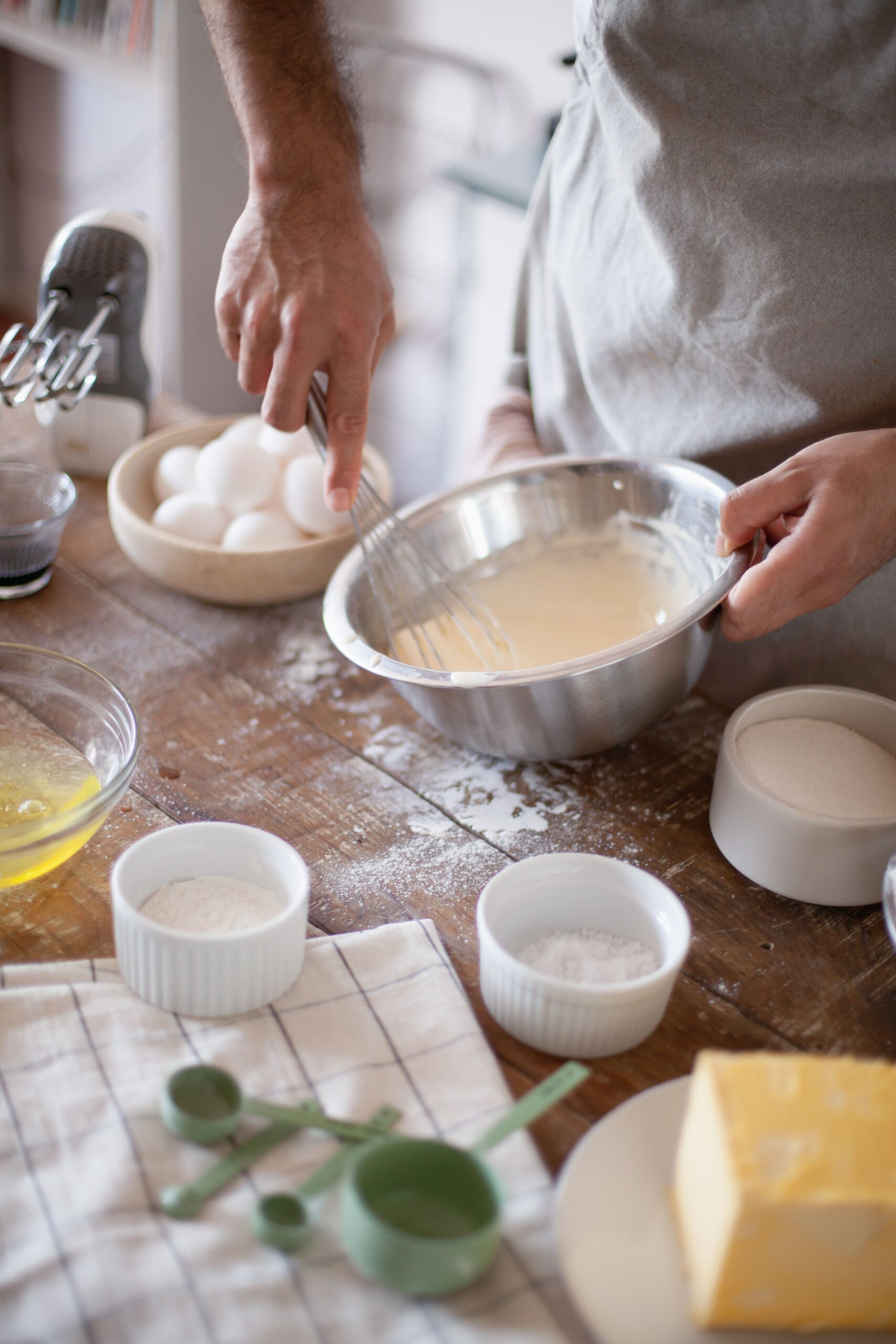 A person whisks cake batter in a metal bowl surrounded by eggs, flour, sugar, and butter