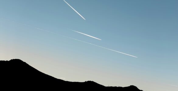 Several white airplane contrails crossing a clear blue sky above a dark mountain ridge.