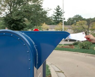 A hand places a stamped envelope into a blue mailbox with an American flag in the background