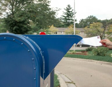 A hand places a stamped envelope into a blue mailbox with an American flag in the background