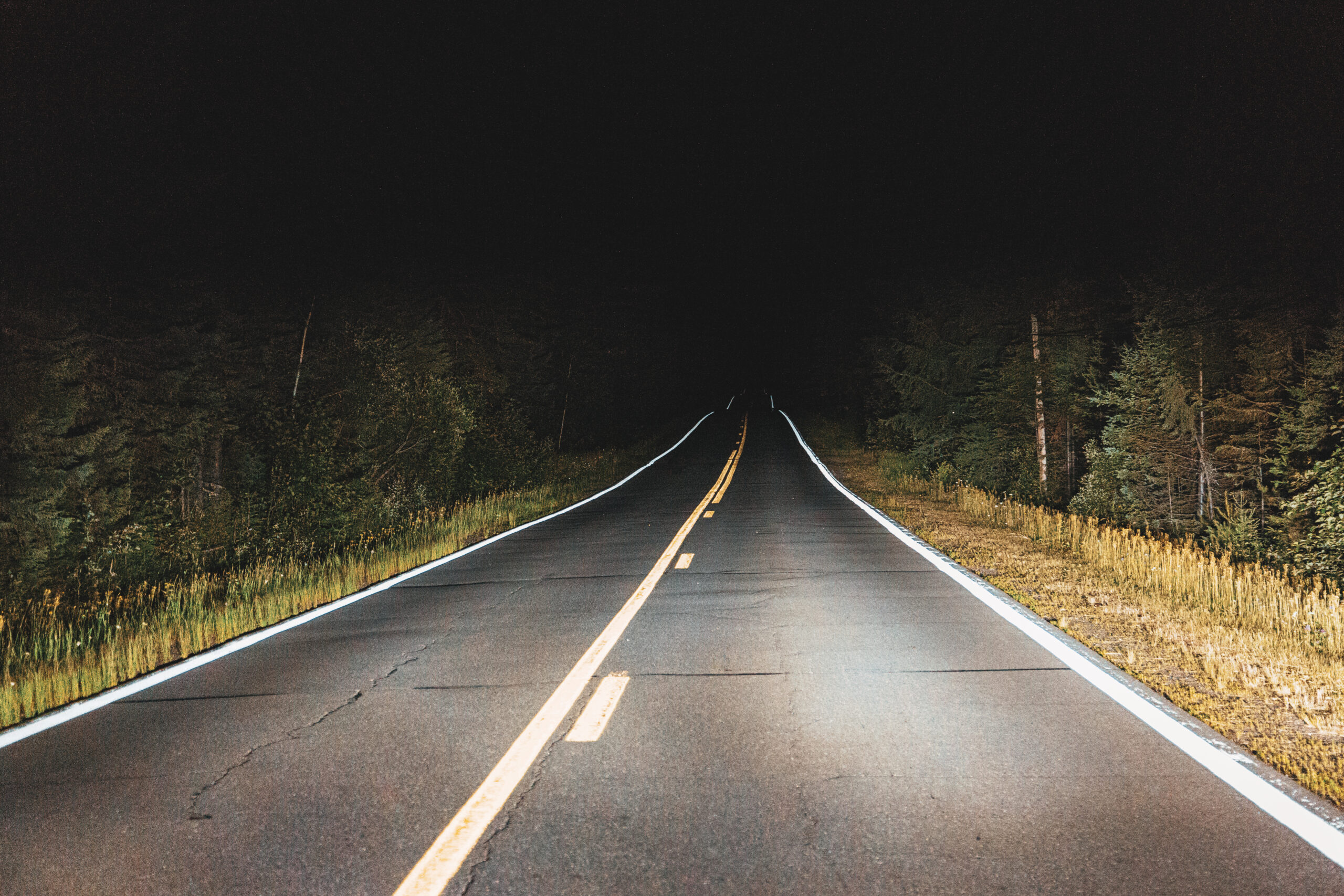 Car headlights illuminating a dark rural road at night, the same light source that makes retroreflective road signs visible.