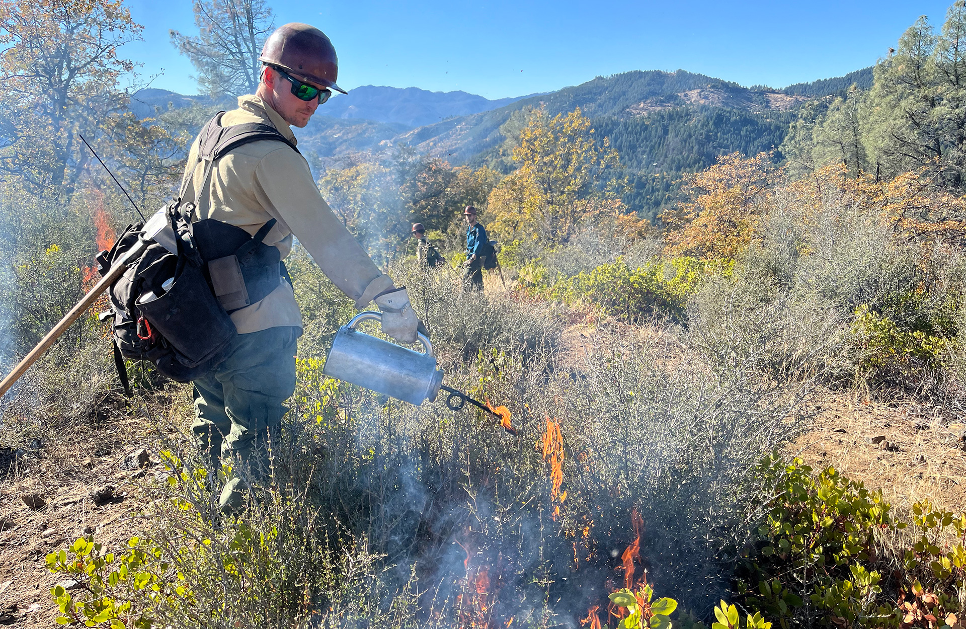 A firefighter uses a drip torch to light a prescribed fire on a forested ridge while other crew members monitor the burn.