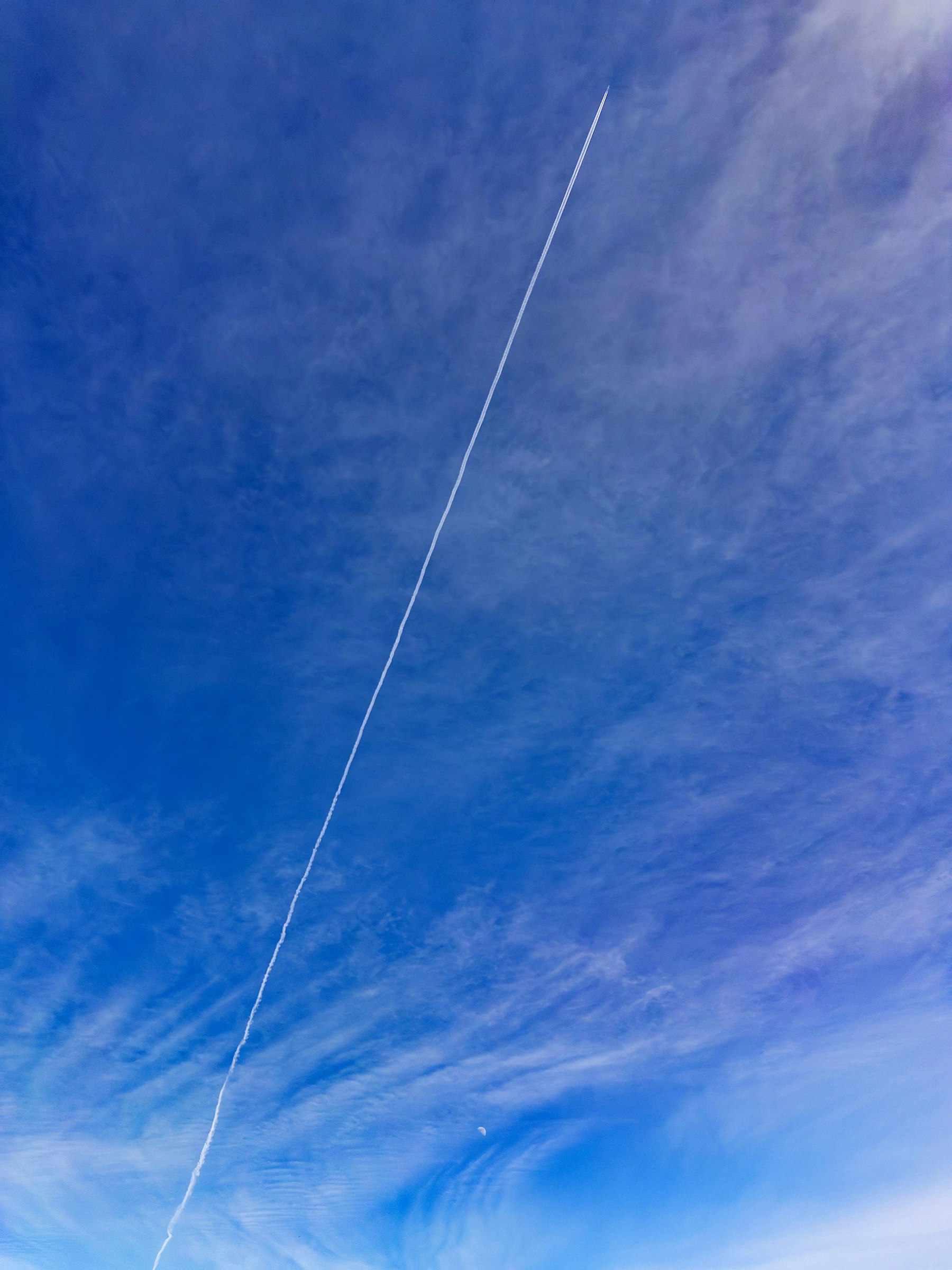 A thin airplane contrail stretching through wispy high clouds in a blue sky.