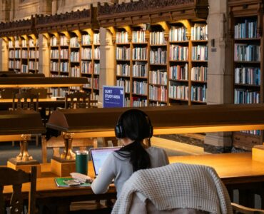 A student works on a laptop in a library while organizing research sources and notes.