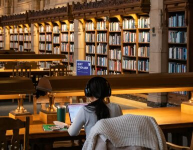 A student works on a laptop in a library while organizing research sources and notes.