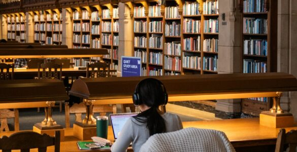 A student works on a laptop in a library while organizing research sources and notes.