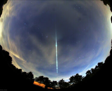 A bright meteor fireball leaves a glowing segmented trail across a fisheye view of the night sky