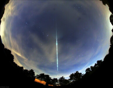 A bright meteor fireball leaves a glowing segmented trail across a fisheye view of the night sky