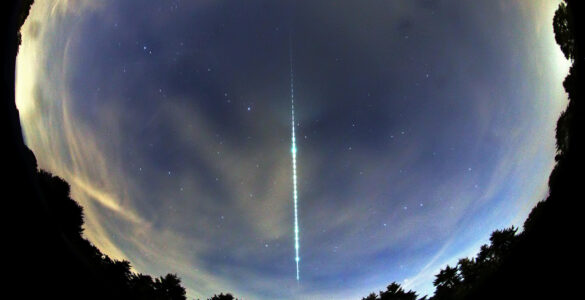 A bright meteor fireball leaves a glowing segmented trail across a fisheye view of the night sky
