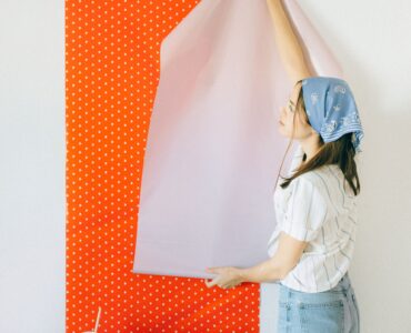 A person applying removable wallpaper to a painted wall, showing the backing peeled away from the adhesive sheet.