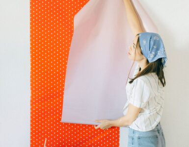 A person applying removable wallpaper to a painted wall, showing the backing peeled away from the adhesive sheet.