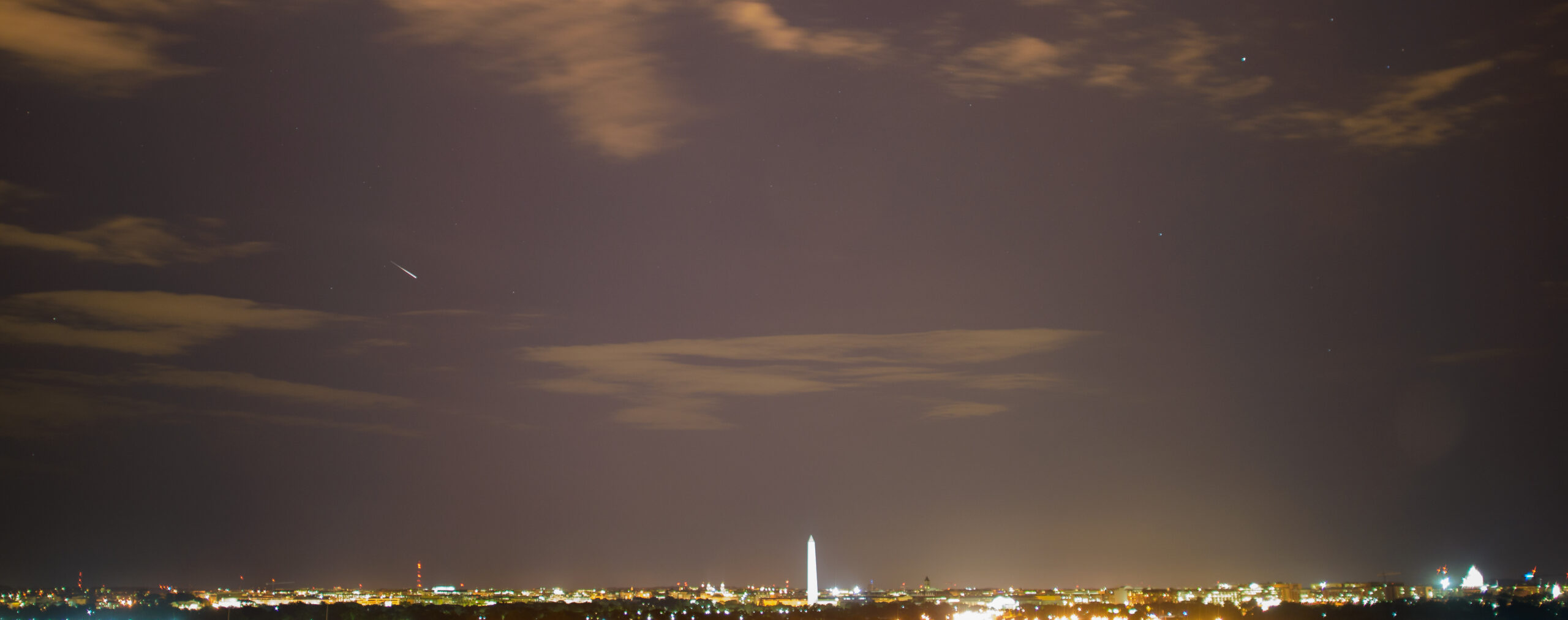 A Perseid meteor creates a bright streak across the night sky above a city horizon