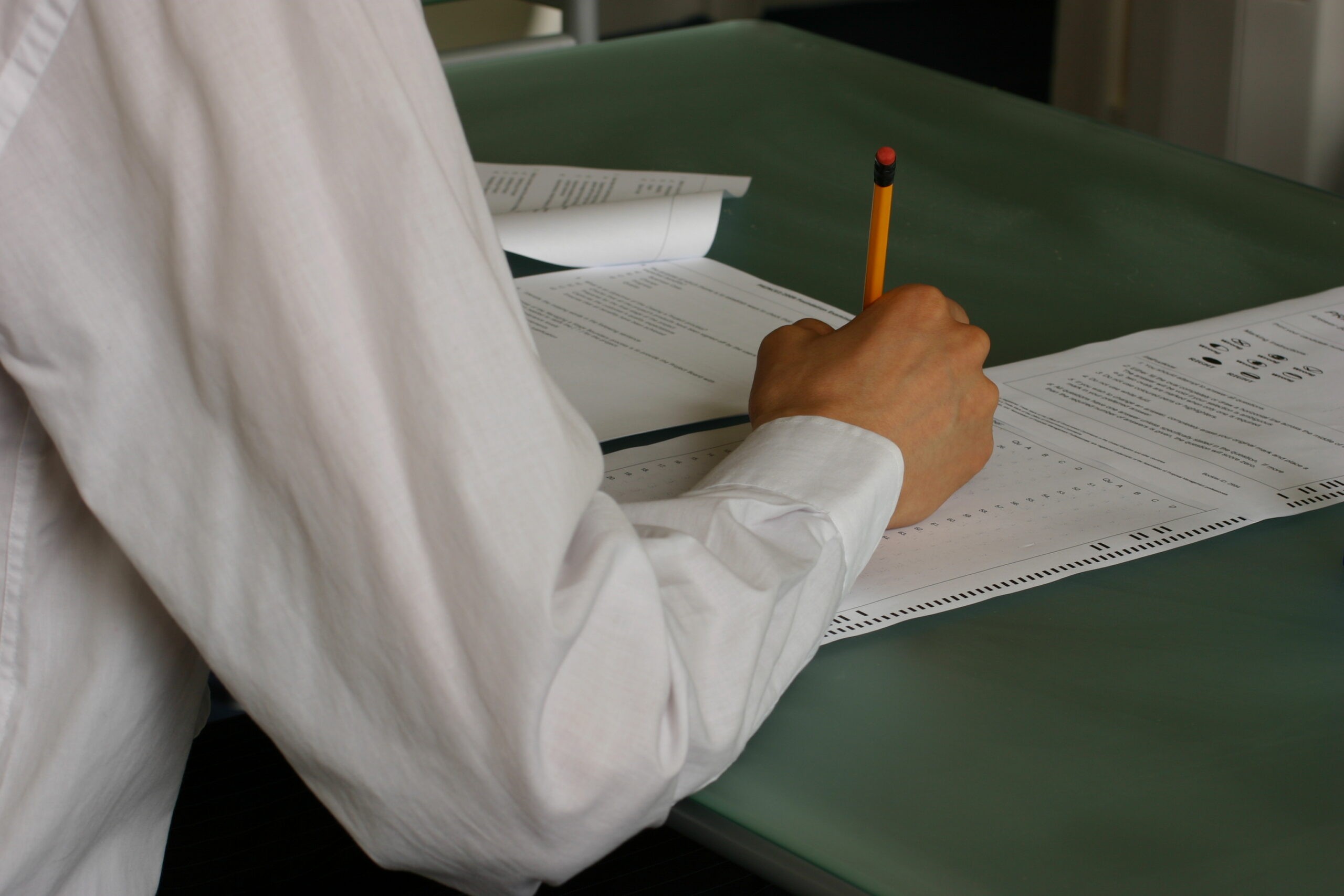 A student writes answers by hand at a classroom desk.