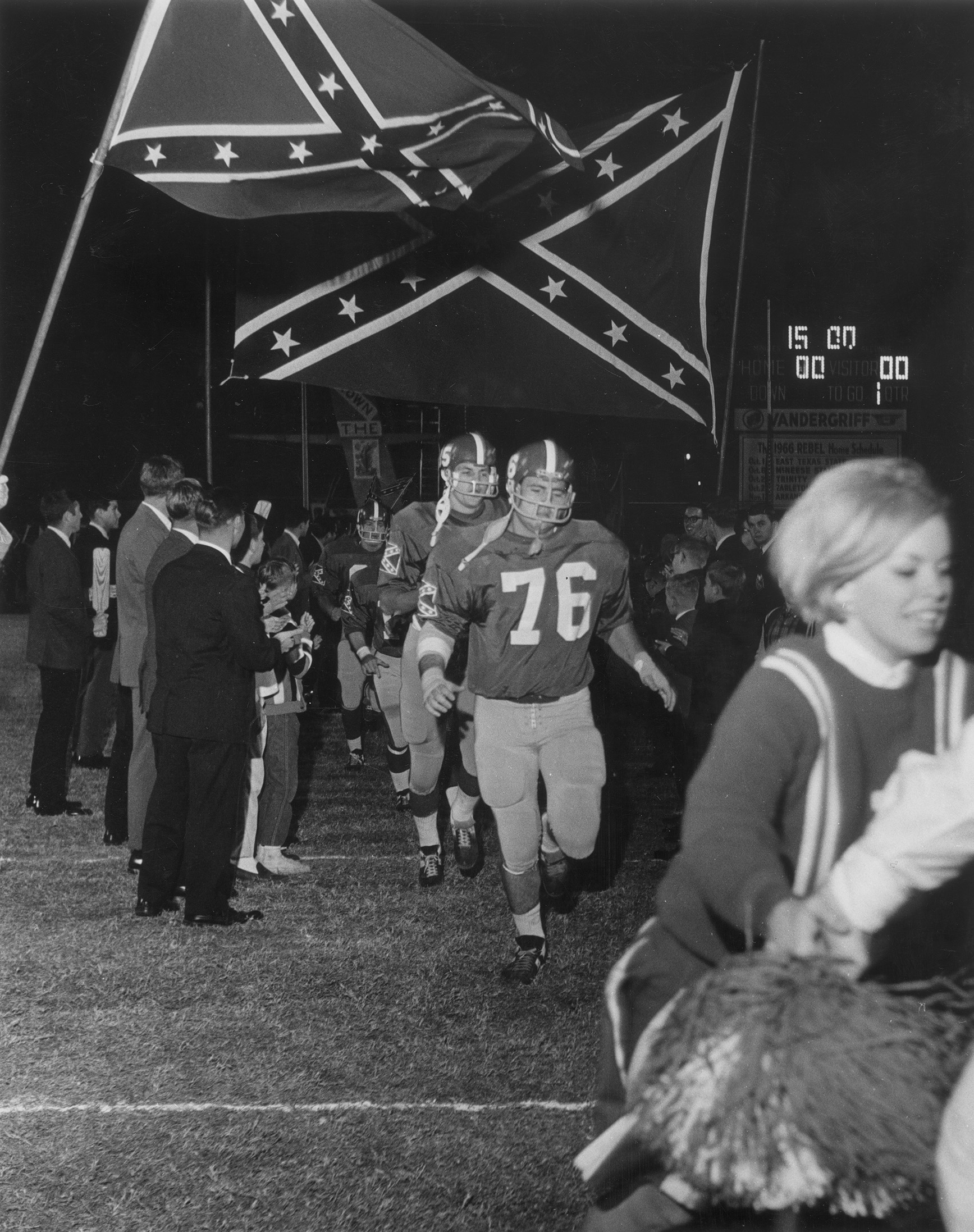 Football players run beneath school flags at Arlington State College, showing the sports setting that shaped many homecoming events.
