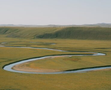 A winding river crosses a broad floodplain, showing bends shaped by erosion and sediment deposition.