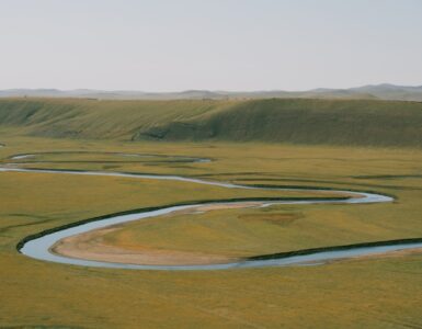 A winding river crosses a broad floodplain, showing bends shaped by erosion and sediment deposition.