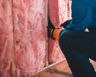 A worker fitting pink insulation between wall studs in a building interior.