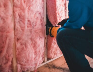 A worker fitting pink insulation between wall studs in a building interior.