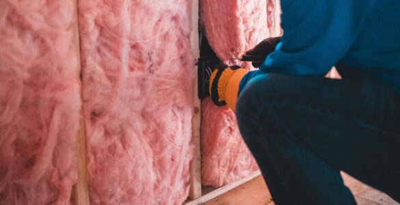 A worker fitting pink insulation between wall studs in a building interior.