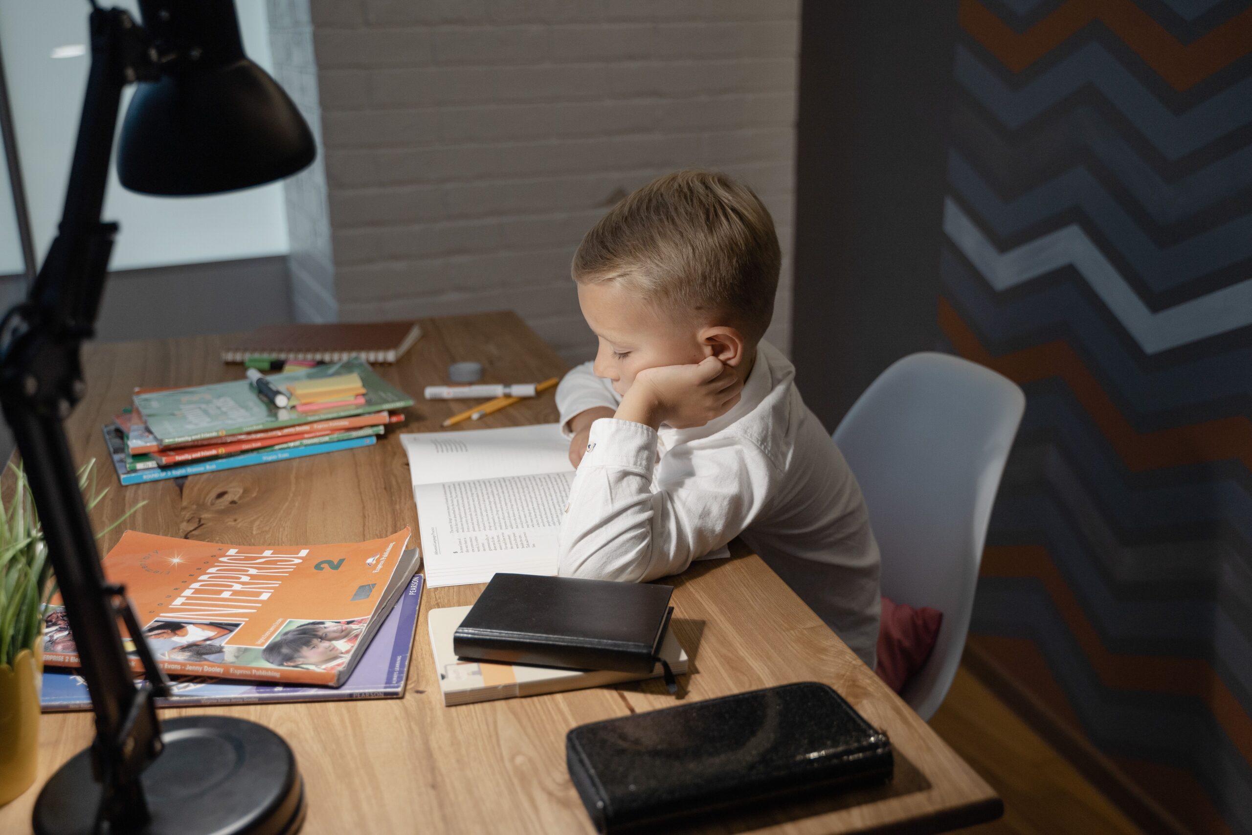 Boy reading a book at a desk with notebooks and a lamp